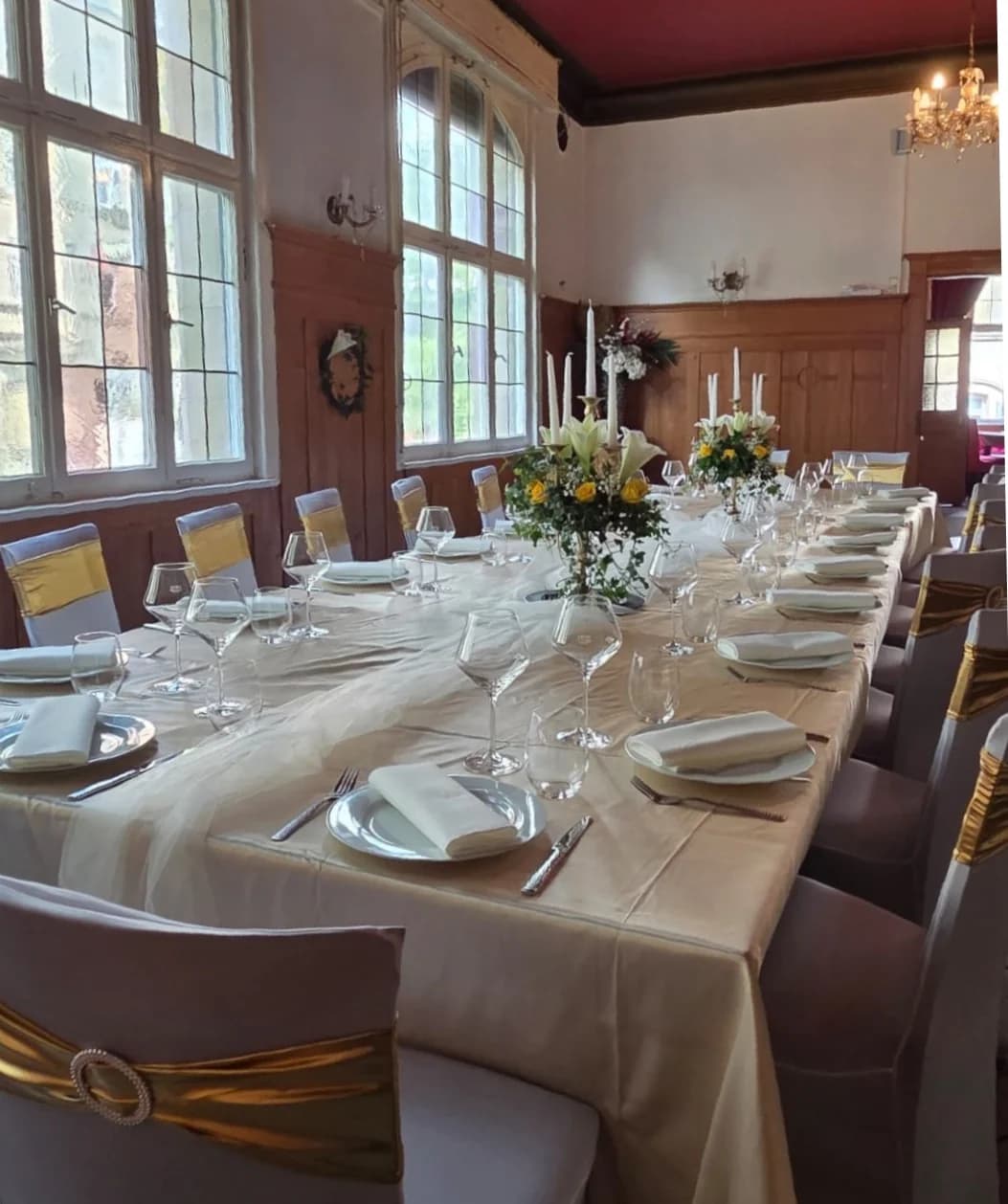 Festlich gedeckte lange Tafel im Goldoni-Saal mit hohen Bogenfenstern, weißen Stuhlhussen mit goldenem Band, Kandelabern mit weißen Kerzen und Tischdeko aus gelben Rosen und Lilien.