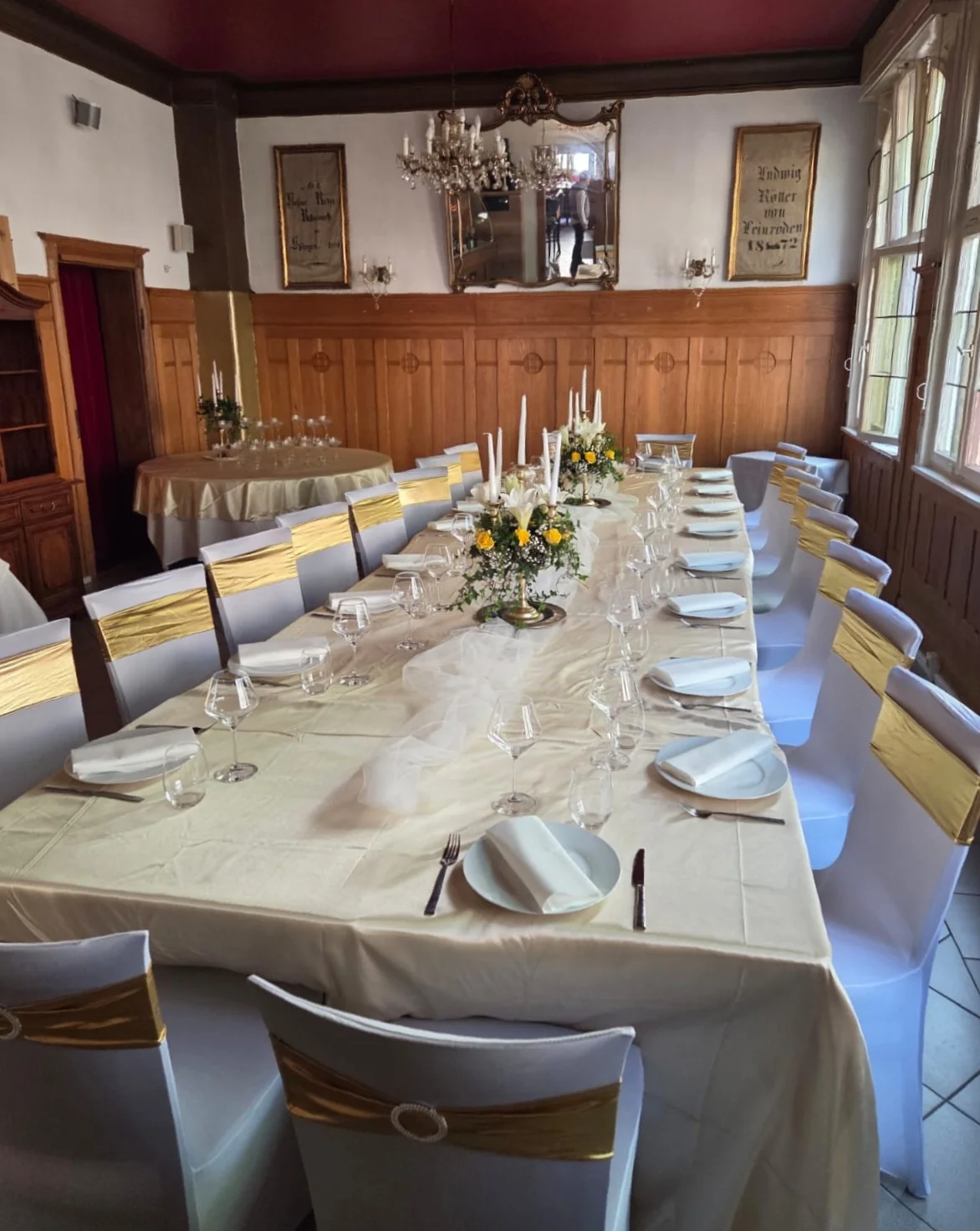 Goldoni-Saal mit historischen Wandbannern (Ludwig Köller von Leinroden, 1872), Spiegel, Holzvertäfelung und festlich gedeckter Tafel mit weißen Stuhlhussen und goldenen Bändern.