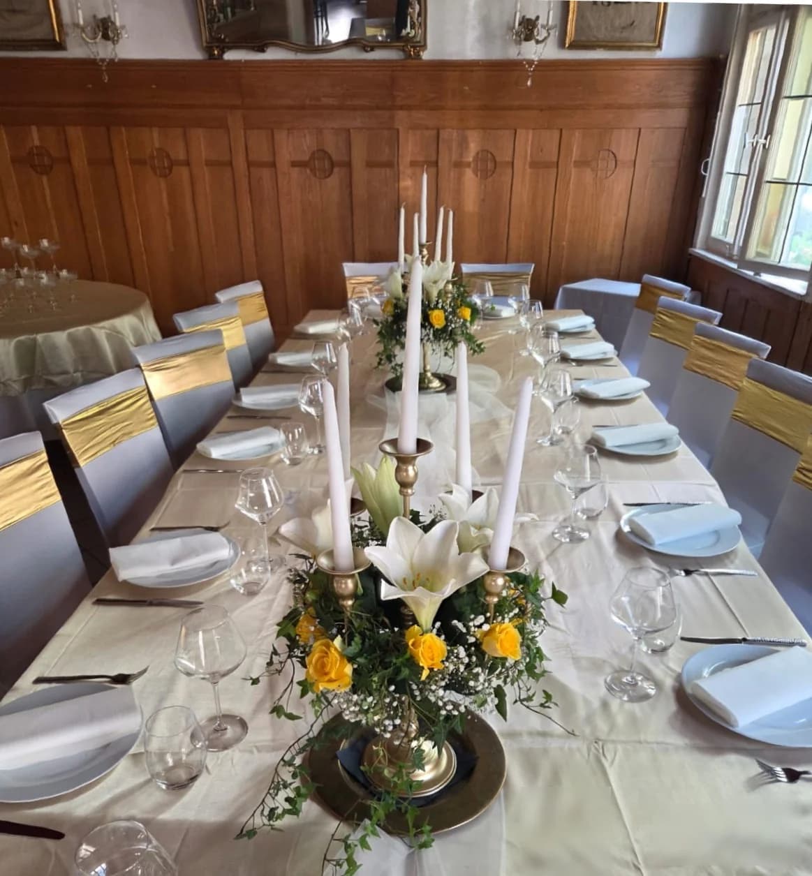 Festlich eingedeckte Tafel von oben — weiße Teller, Kristallgläser, Stuhlhussen mit goldenem Satinband, Tischdeko mit Kandelaber und Blumen.